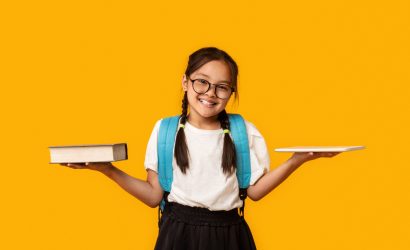 Gadgets Vs Books. Asian School Girl Comparing Digital Tablet And Book Standing Over Yellow Studio Background.